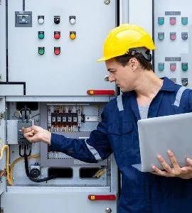 An engineer inspecting a distribution board with a laptop in hand.